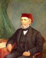 White man with grey beard, wearing suit and red fez, seated in a wooden armchair and resting his elbow near books on a table.